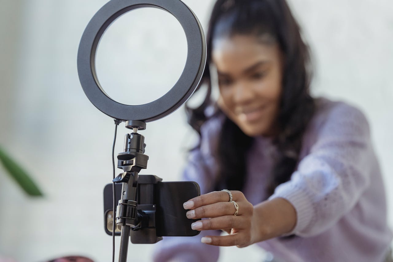 creative-03 Cheerful young African American female blogger in stylish sweater smiling while setting up camera of smartphone attached to tripod with ring light before recording vlog